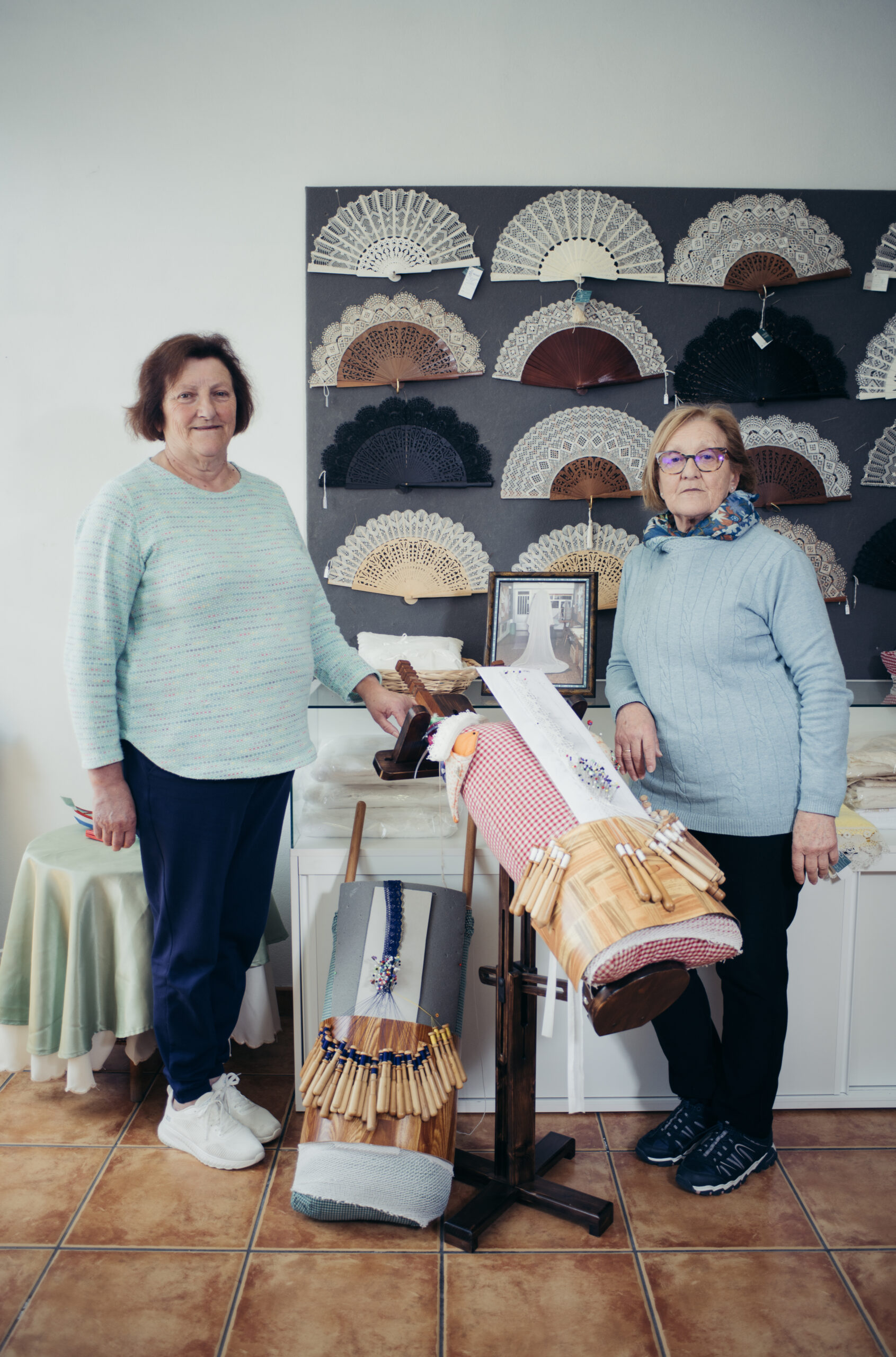 Dos mujeres tejedoras posan junto a sus obras de encaje de bolillos, con una pared de fondo decorada con abanicos artesanales de encaje.