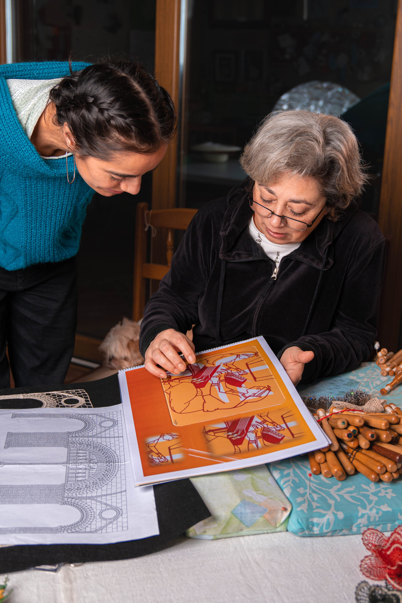 Ana Rosa Lista y una mujer revisando un diseño gráfico de encaje de bolillos en una mesa con herramientas de trabajo.