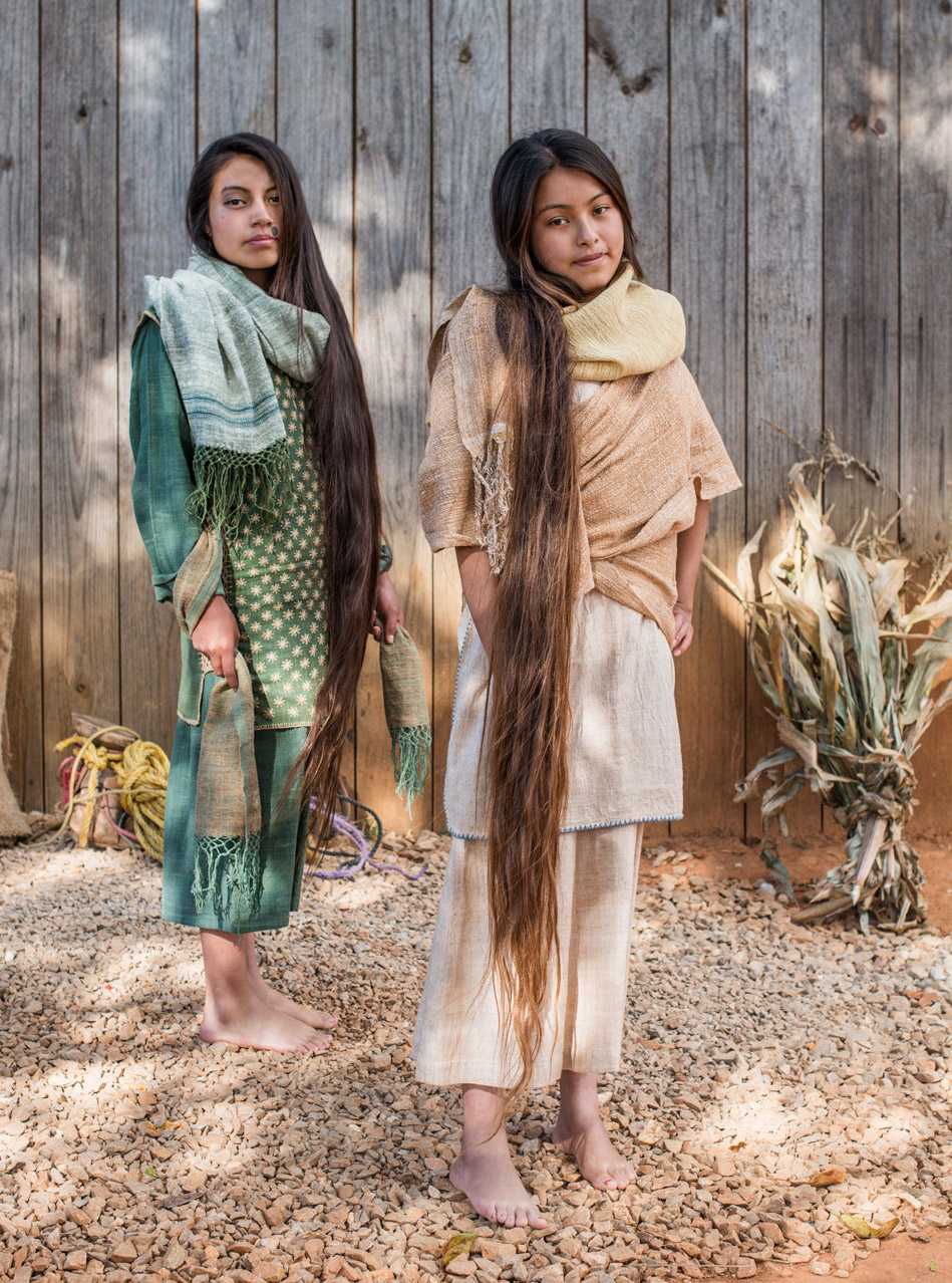 Dos artesanas de la comunidad mexicana Khadi Oaxaca, vestidas con prendas tradicionales hechas a mano, posan frente a una pared de madera, destacando la riqueza de la artesanía textil.